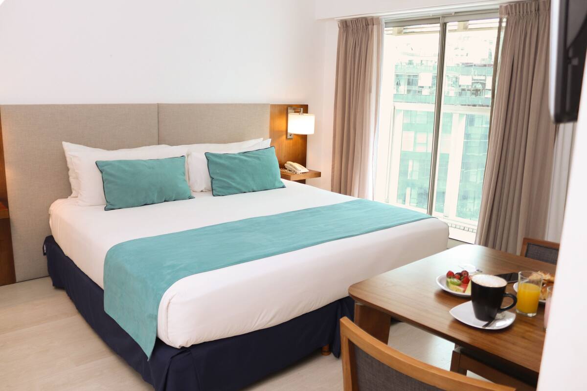 A hotel room with a bed, a bedside table with a lamp, a dining table with breakfast items, and a view of buildings through the window.