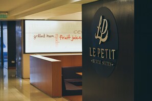 Un espace d’accueil au design moderne, comprenant un comptoir de réception et un coin salon. Un panneau mural affiche les mentions « jambon grillé, jus de fruits » et « LE PETIT RENTAL SUITES ».