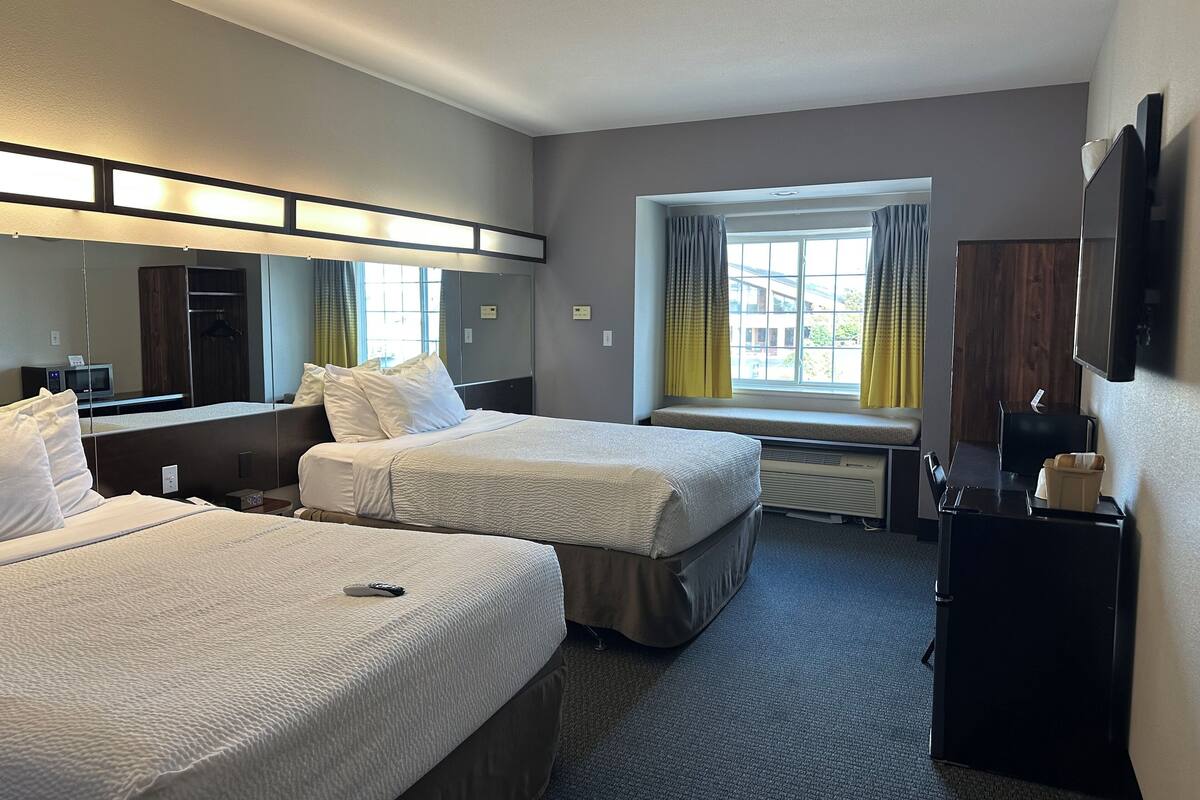 Ein Hotelzimmer mit zwei Betten, einem großen Spiegel, einem Fenster mit Vorhängen, einem Flachbildfernseher und einem Schreibtisch mit Telefon und Taschentuchbox.