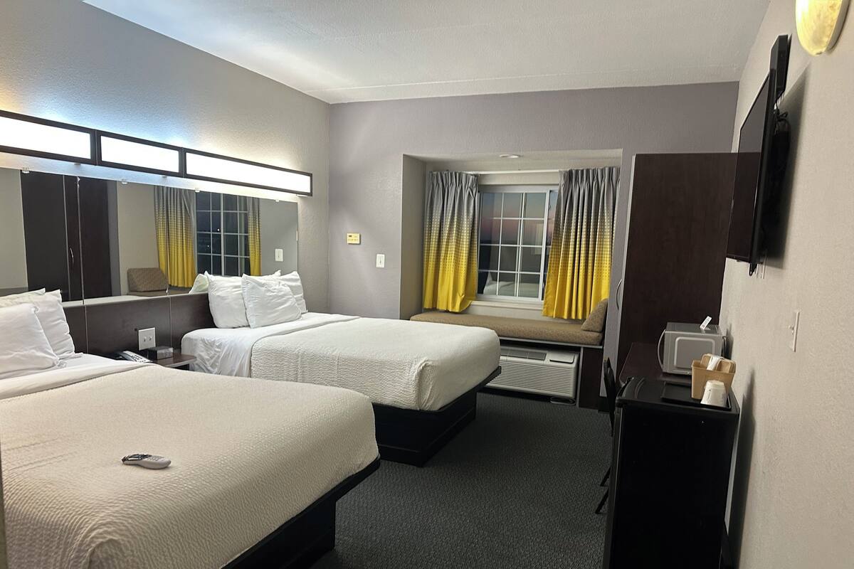 Ein Hotelzimmer mit zwei Betten, einem Schreibtisch, einem Stuhl, einem Spiegel und einem Fenster mit Vorhängen.