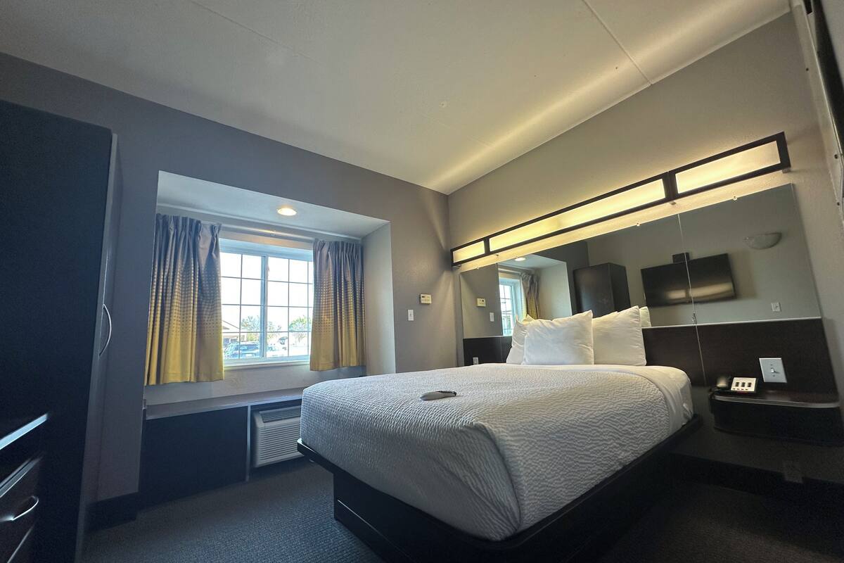 Ein Hotelzimmer mit einem großen Bett, einem Nachttisch mit Lampe, einem Fenster mit Vorhängen und einem an der Wand befestigten Flachbildfernseher.