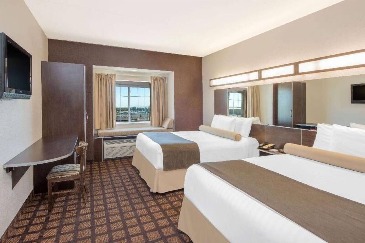 Ein Hotelzimmer mit zwei Betten, einem Schreibtisch, einem Stuhl, einem Fernseher, einem Fenster mit Vorhängen und Blick auf die Stadt.