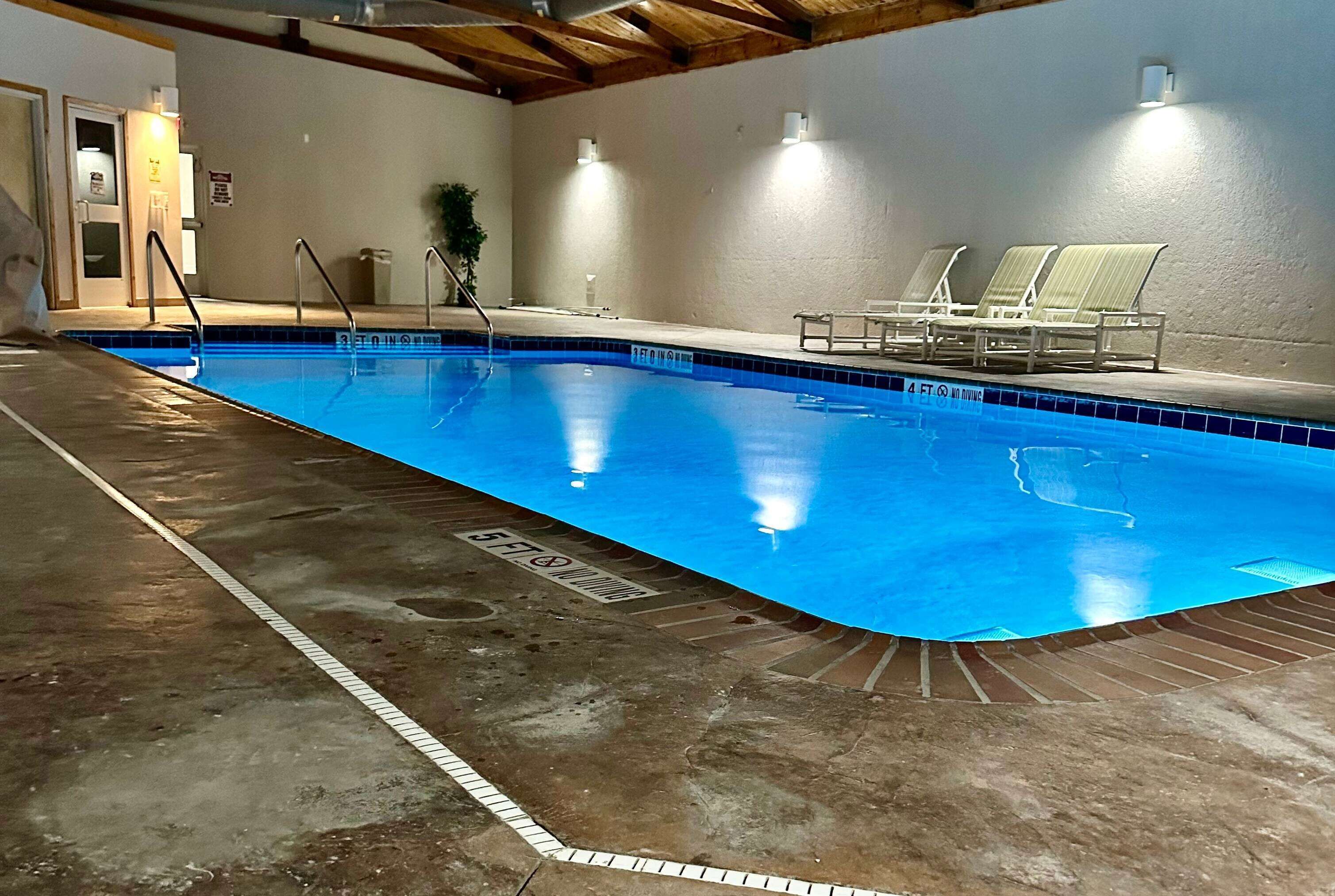 An indoor swimming pool with clear lane markings, a tiled floor, and lounge chairs along the side.