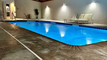 An indoor swimming pool with clear lane markings, a tiled floor, and lounge chairs along the side.