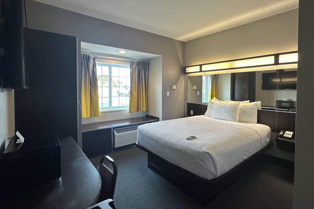 Ein Hotelzimmer mit einem Bett, einem Schreibtisch, einem Sessel, einem Fenster mit Vorhängen, einem Spiegel, einem Fernseher und einem Telefon auf dem Nachttisch.