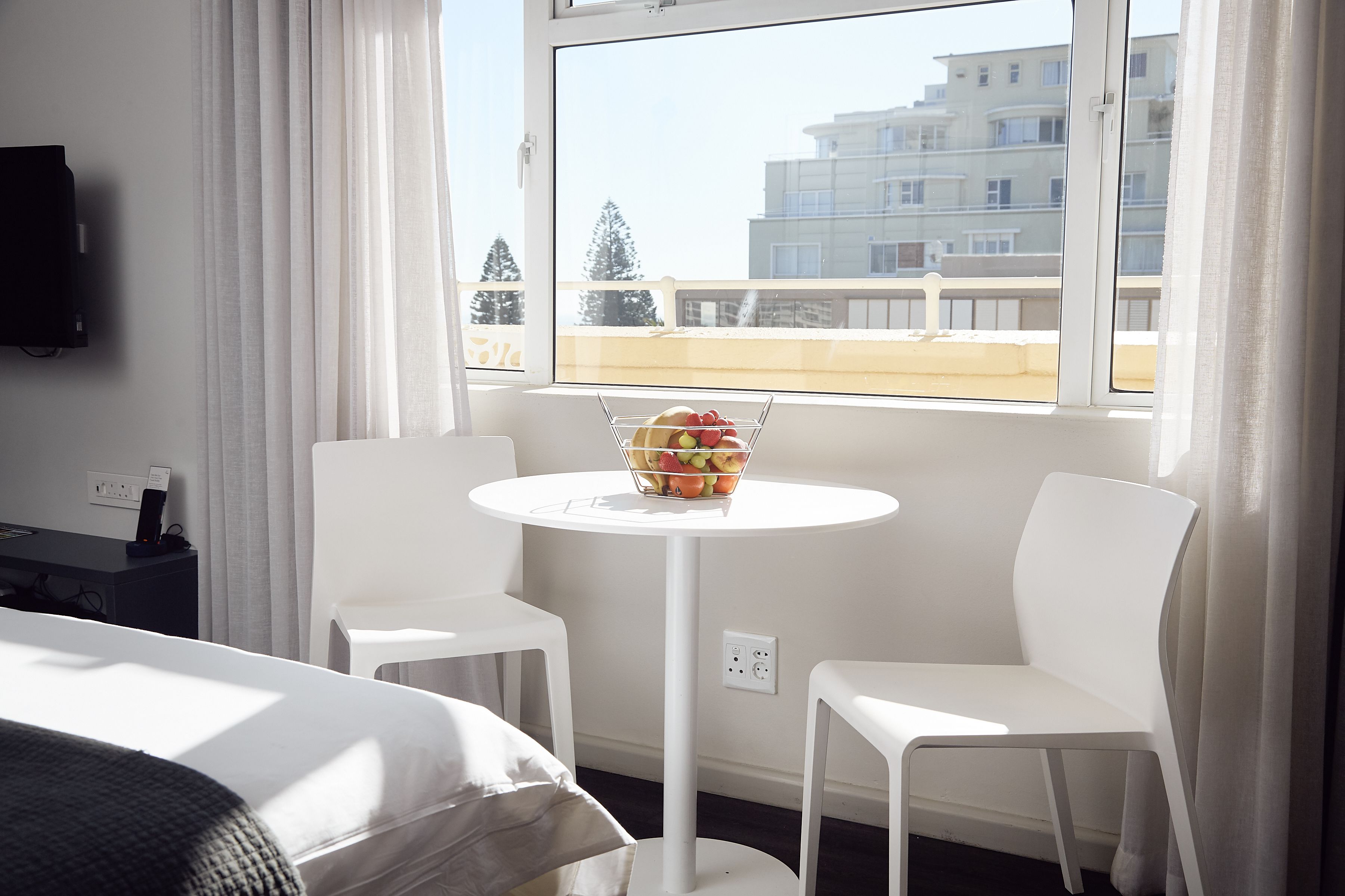 A hotel room with a bed, a white table with a fruit bowl, two white chairs, a TV on the wall, and a window with a view of buildings and trees.