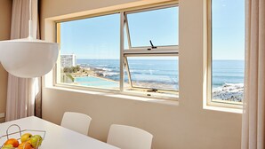 A room with a large window overlooking a beach and ocean, a dining table with chairs, and a fruit basket. - First Group Riviera Suites (Cape Town)