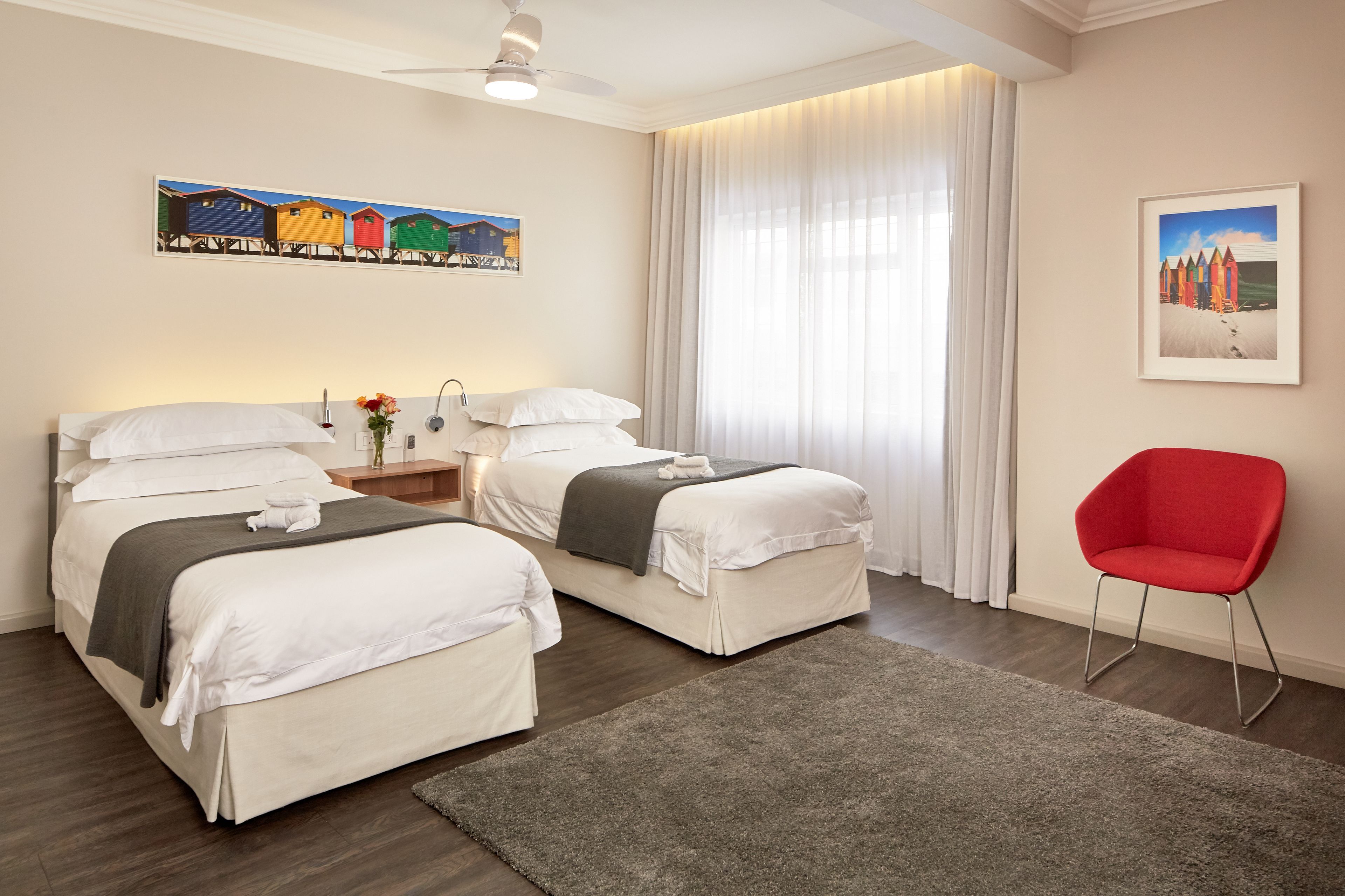 Habitación de hotel con dos camas, una silla roja, un cuadro con cabañas en la playa y un cuadro de un paisaje nevado.