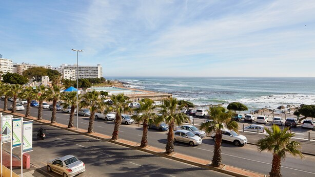 A coastal road lined with palm trees and parked cars, leading to a beach. - First Group Riviera Suites (Cape Town)