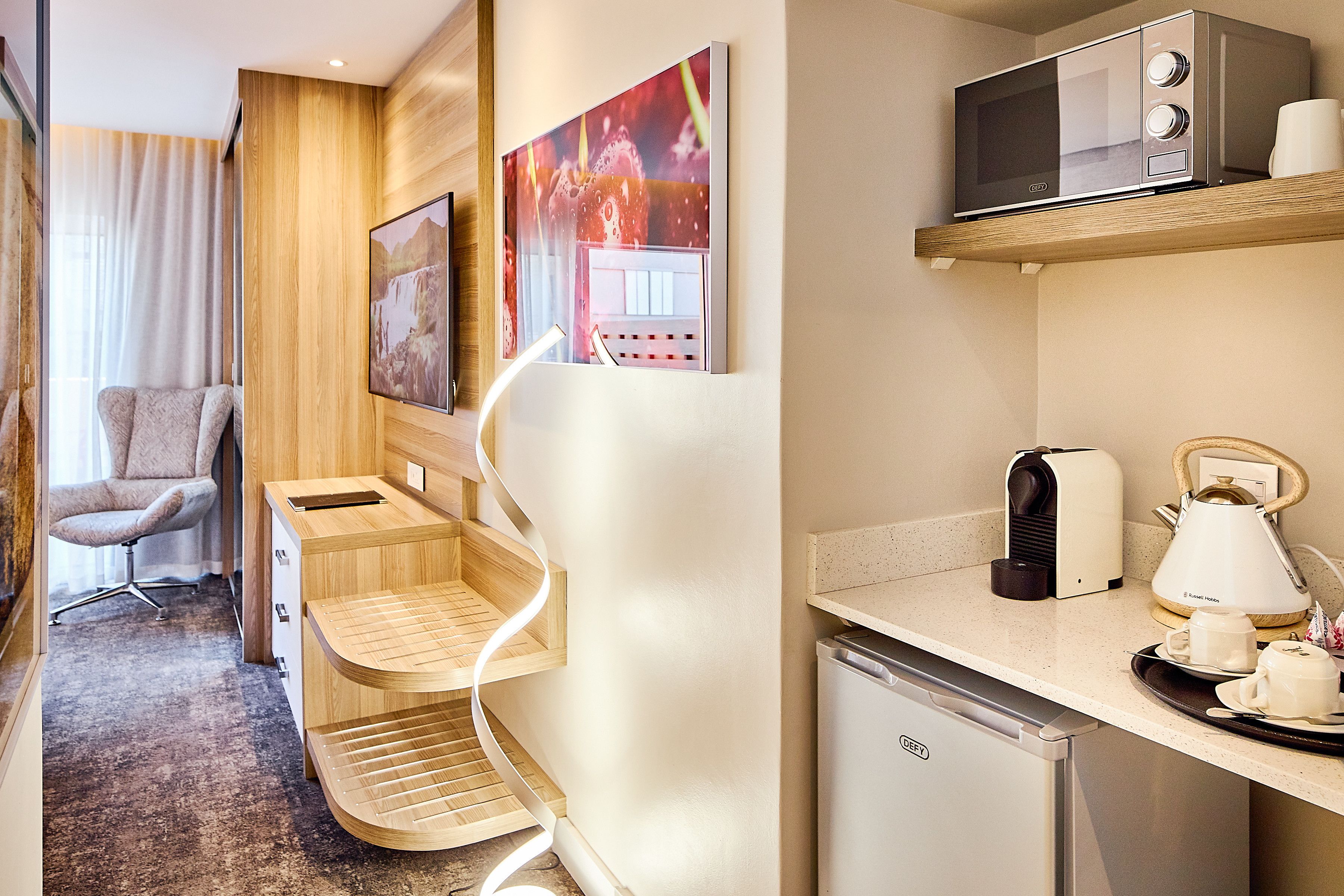 A modern hotel room with a wooden desk, a microwave, a kettle, and a small refrigerator.