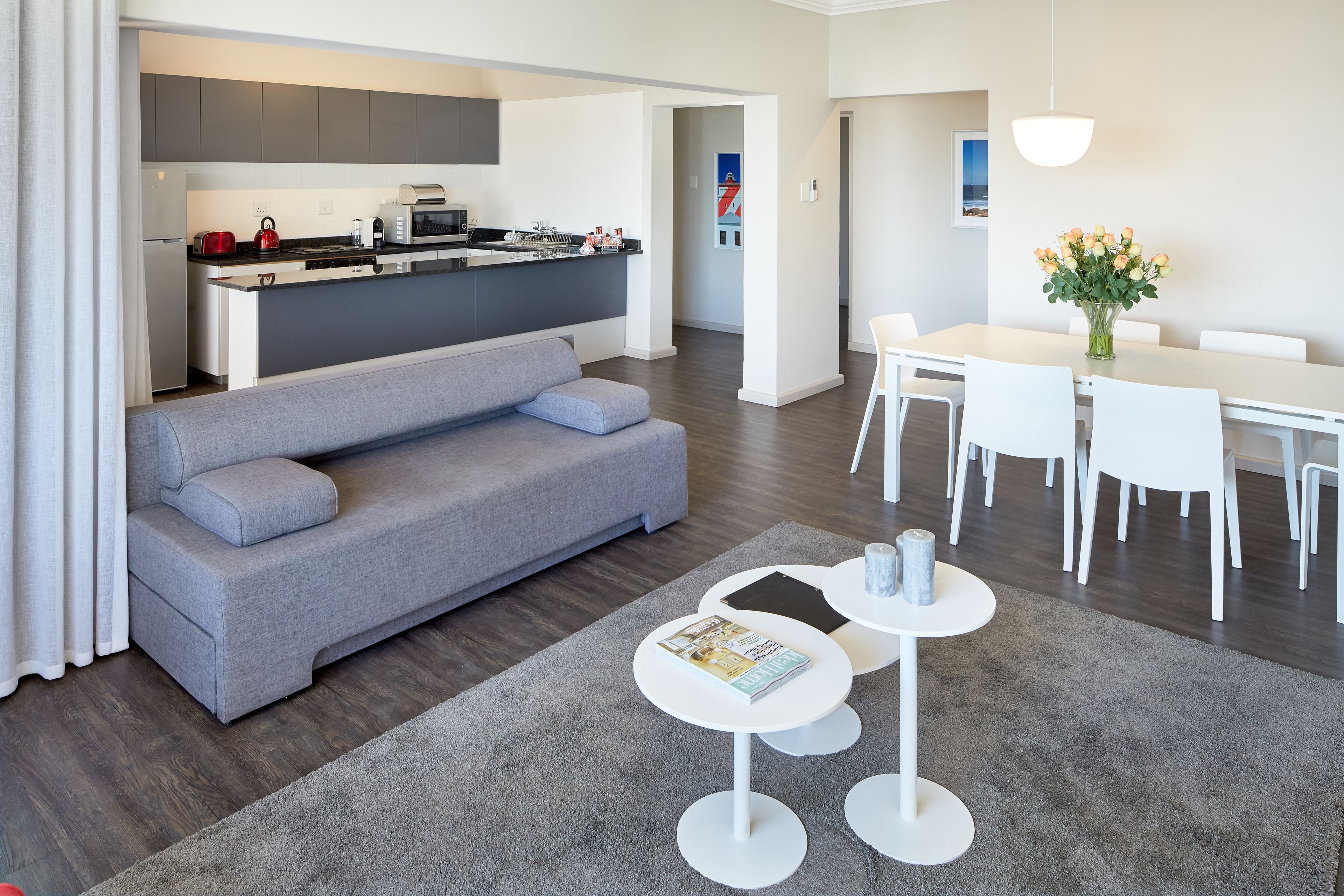 A modern living room with a grey sofa, a dining table with white chairs, and a kitchen in the background.