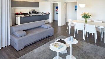 A modern living room with a grey sofa, a dining table with white chairs, and a kitchen in the background.