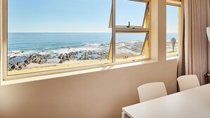A room with a large window overlooking a rocky beach and ocean. There is a table and two chairs in the room. - First Group Riviera Suites (Cape Town)