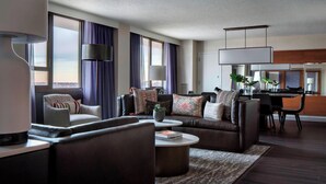 A modern living room with a dark leather sofa, a striped armchair, and a coffee table. There is a dining area with a table and chairs, and a large window with curtains. - New York LaGuardia Airport Marriott (East Elmhurst)