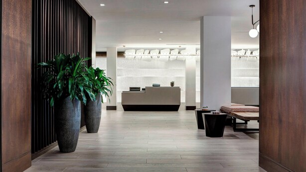 A modern reception area with a wooden reception desk, large potted plants, and a seating area with a bench and table. - New York LaGuardia Airport Marriott (East Elmhurst)