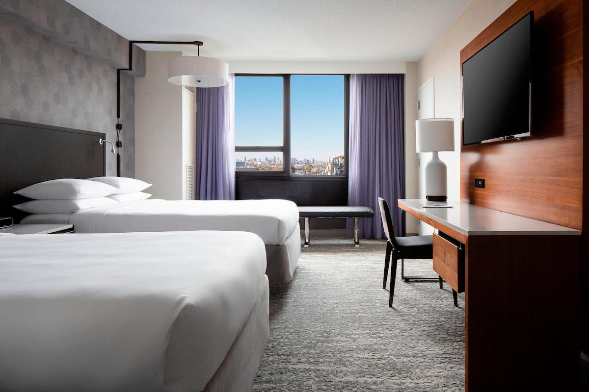 A hotel room with two beds, a desk, a chair, a TV, and a window with a city view.