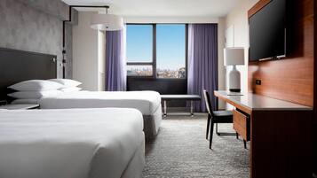 A hotel room with two beds, a desk, a chair, a TV, and a window with a city view.