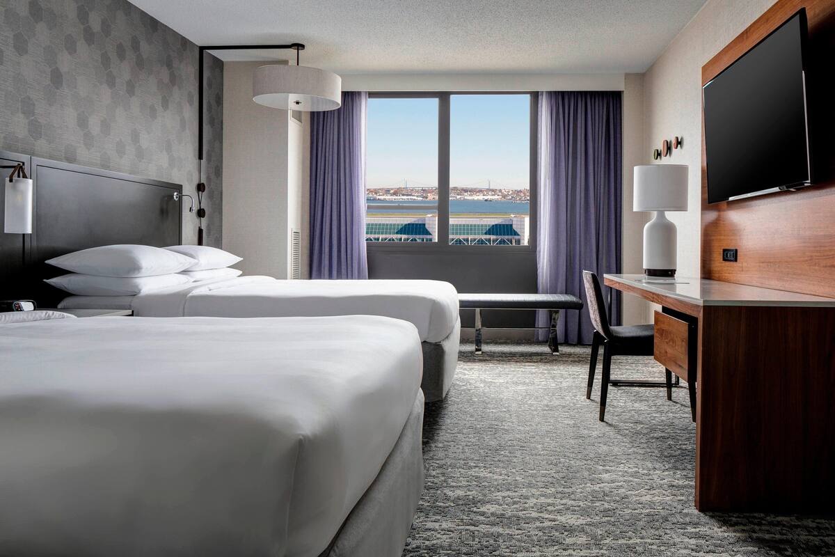 A hotel room with two beds, a desk, a chair, a TV, and a window with a view of a cityscape.