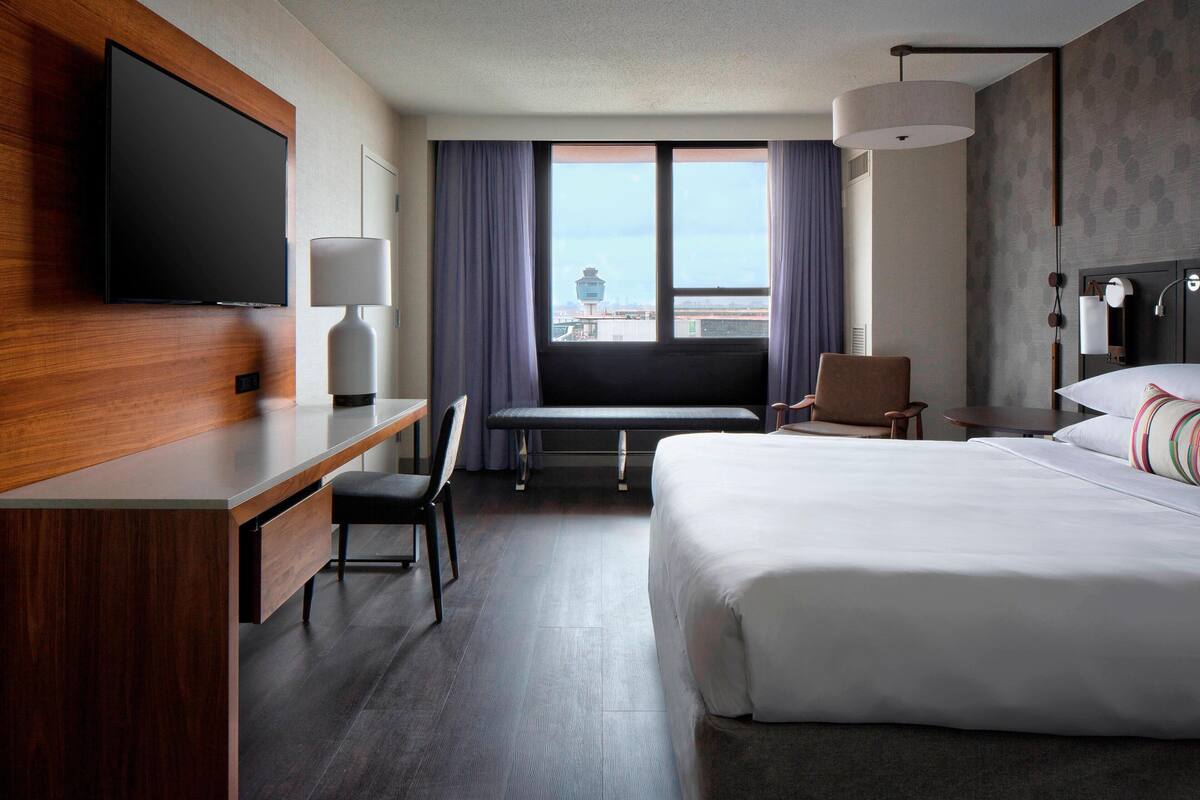 A hotel room with a bed, desk, chair, TV, lamp, and large windows with a view.