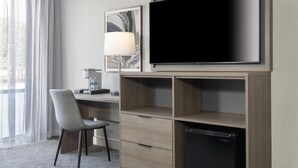 Habitación de hotel con escritorio, una silla, televisor, lámpara y minibar.