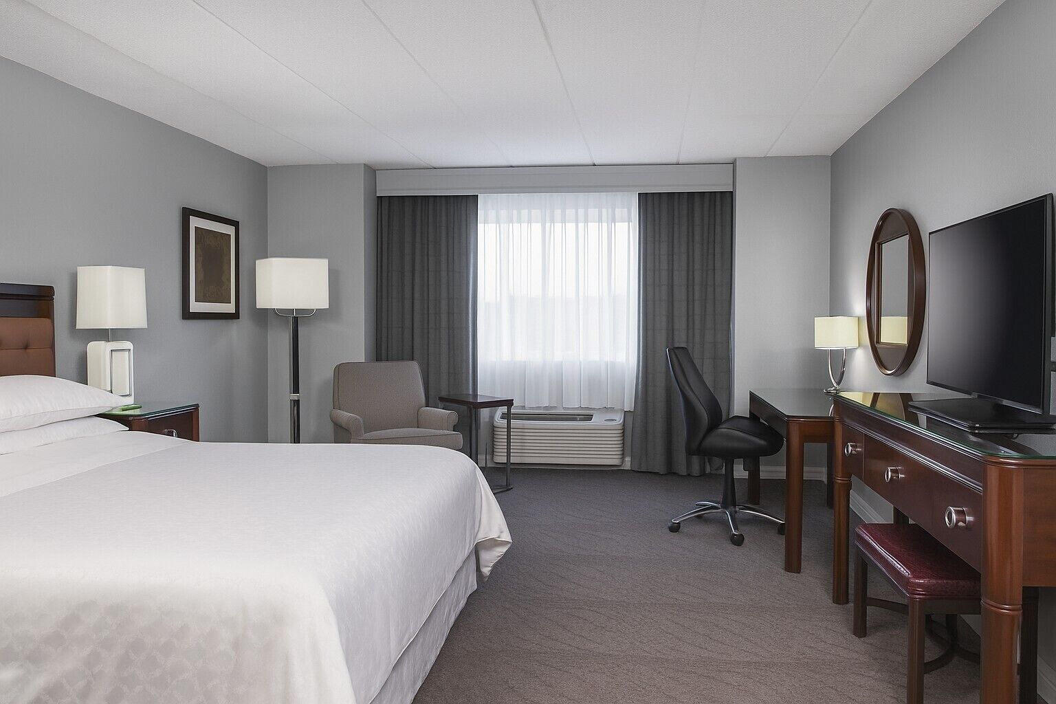 A hotel room with a bed, a desk with a TV, a chair, a lamp, and an air conditioning unit.