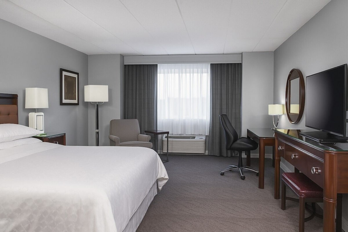 Una habitación de hotel con una cama, un escritorio con televisor, una silla, una lámpara y un equipo de aire acondicionado.