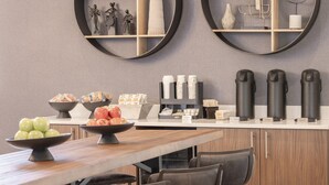 Una cocina moderna con una mesa de madera, sillas y diversos alimentos, como manzanas y pasteles. Hay cafeteras y una estación de café con tazas y espumador de leche.