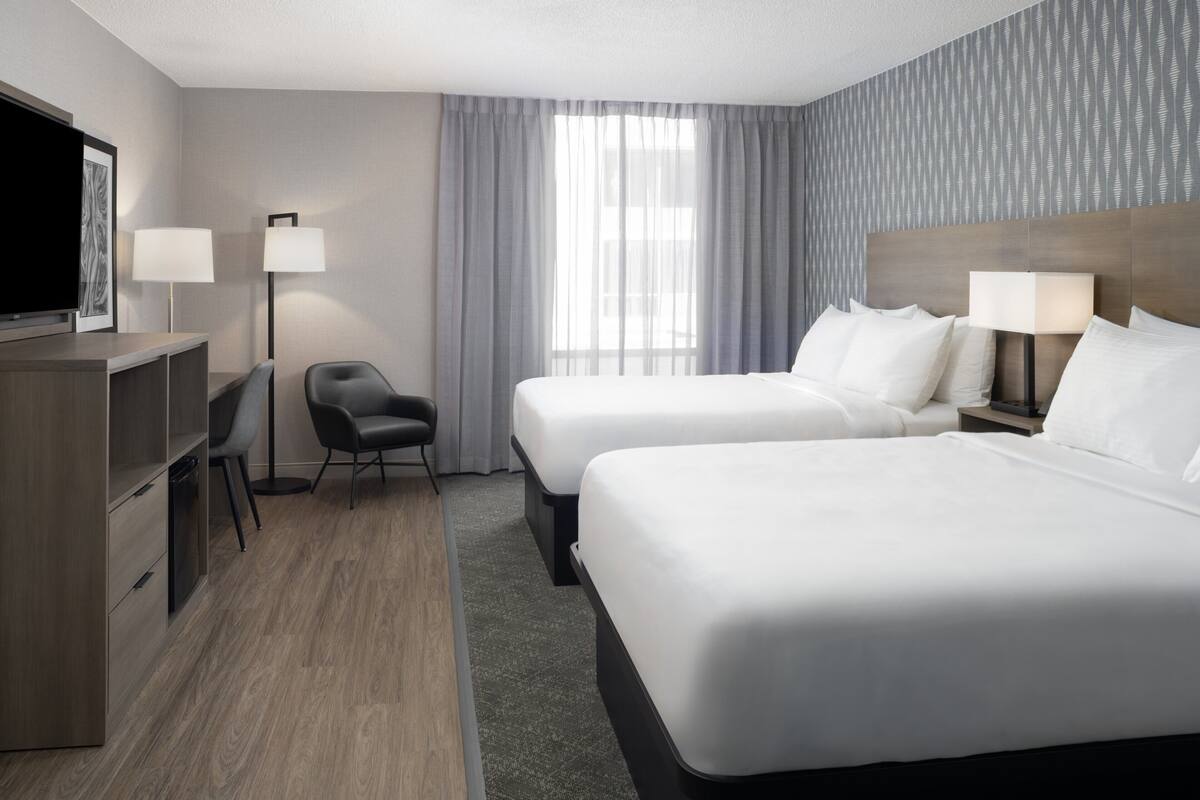 A hotel room with two beds, a desk, a chair, a TV, and a window with curtains.