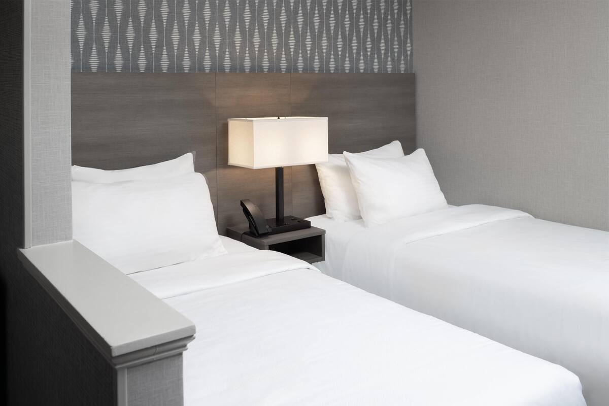 A hotel room with two single beds, a nightstand with a lamp, and a patterned wallpaper on the wall.
