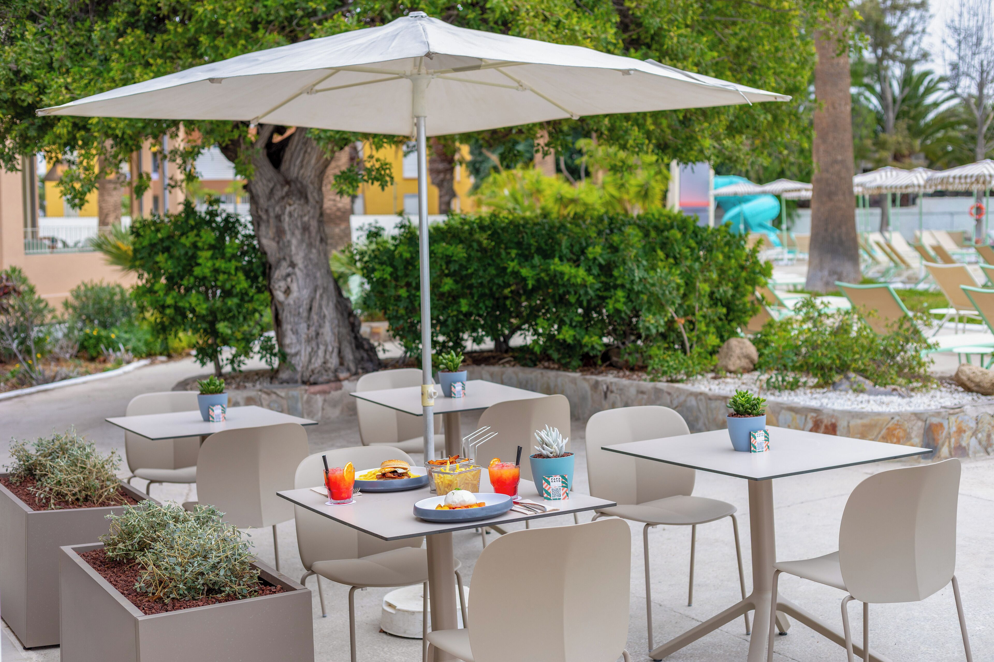 Outdoor seating with tables, chairs, and umbrellas, surrounded by greenery and a pool area.