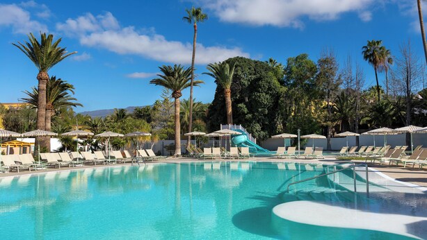 A swimming pool with a water slide, surrounded by palm trees and lounge chairs. - Sol Arona Tenerife (Arona)