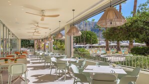 A sunlit outdoor dining area with tables and chairs, ceiling fans, and a view of a pool and palm trees. - Sol Arona Tenerife (Arona)