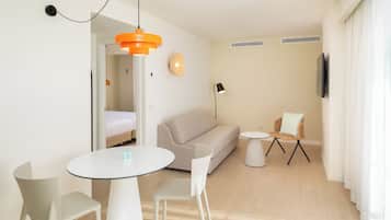 Un salon moderne comprenant un canapé, une table à manger avec des chaises, un téléviseur fixé au mur et une petite table d’appoint avec une lampe.