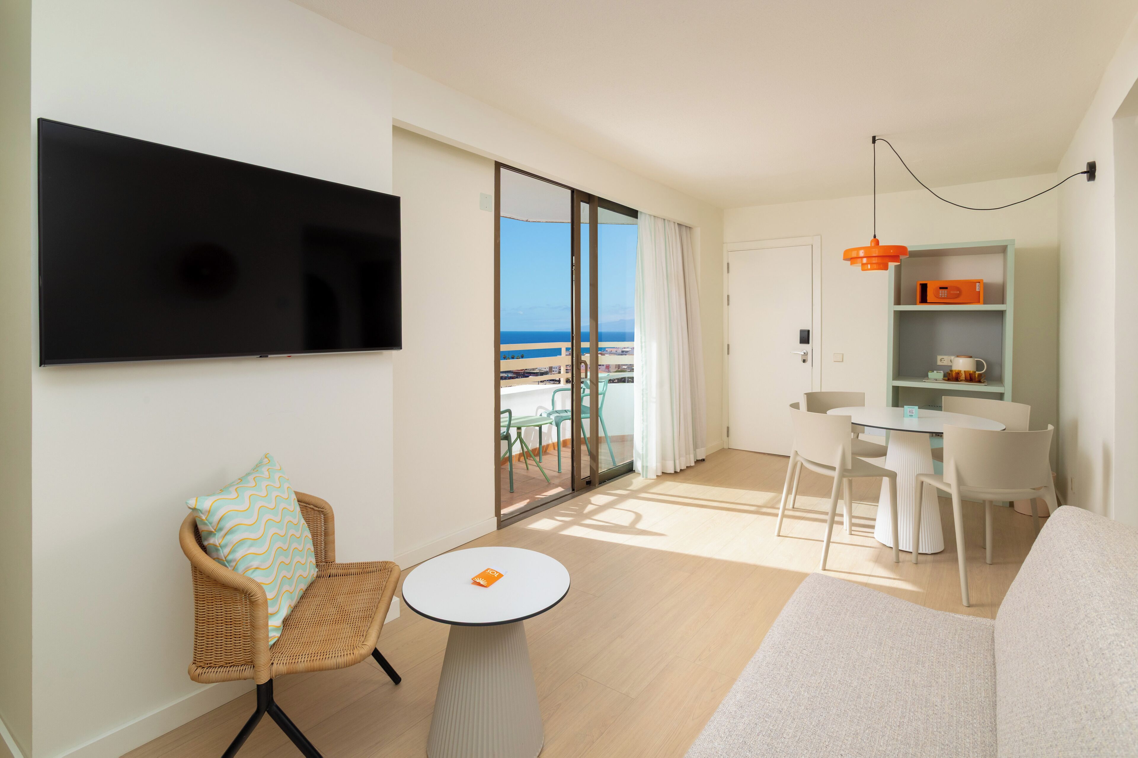 A modern hotel room with a balcony, a flat-screen TV, a dining area, and a sofa.