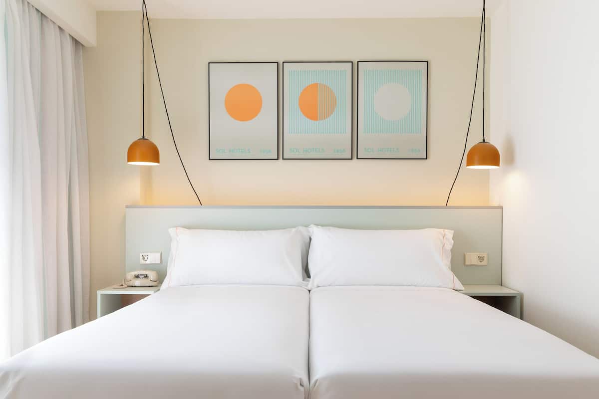 Un lit double avec du linge de lit blanc, deux tables de chevet, un téléphone et trois œuvres d’art encadrées accrochées au mur.