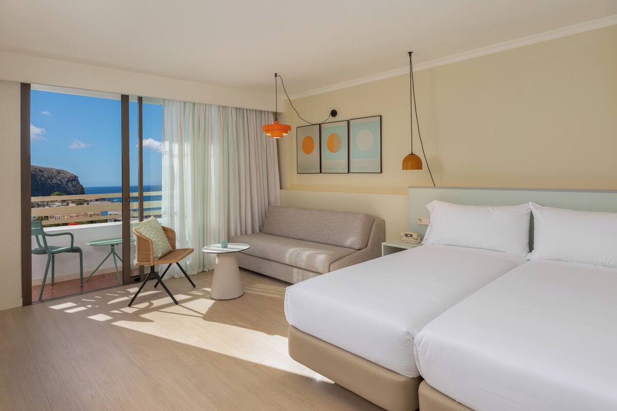 Une chambre d’hôtel avec un grand lit, un canapé, une petite table et une chaise. La fenêtre offre une vue sur la plage.