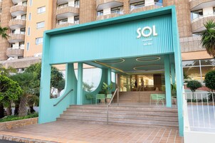 Entrada de un hotel con fachada azul aguamarina, una escalera que conduce al vestíbulo y un letrero que dice 'SOL'.