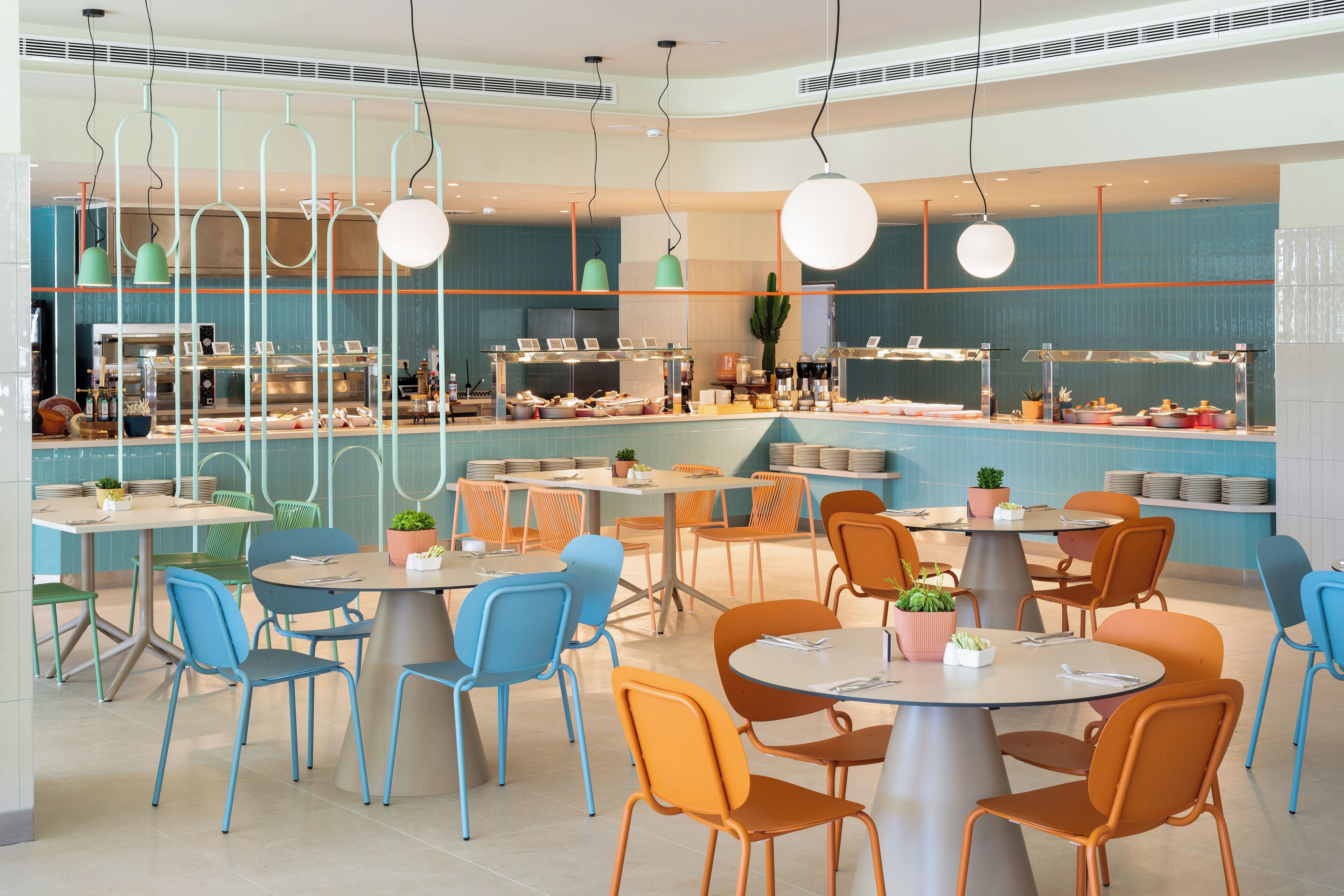 Un restaurante moderno con mesas redondas, sillas anaranjadas y azules, y una barra con exhibición de alimentos.