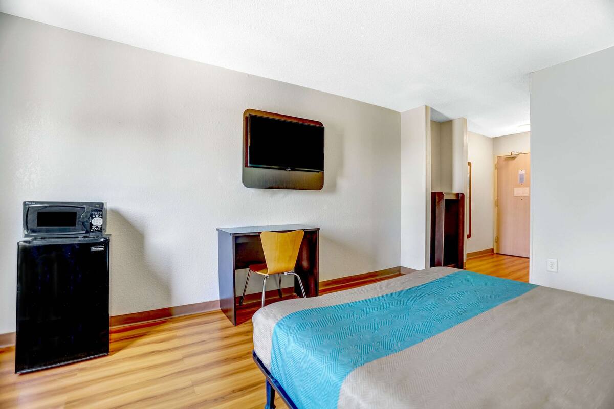A hotel room with a bed, a desk with a chair, a microwave, and a television on the wall.