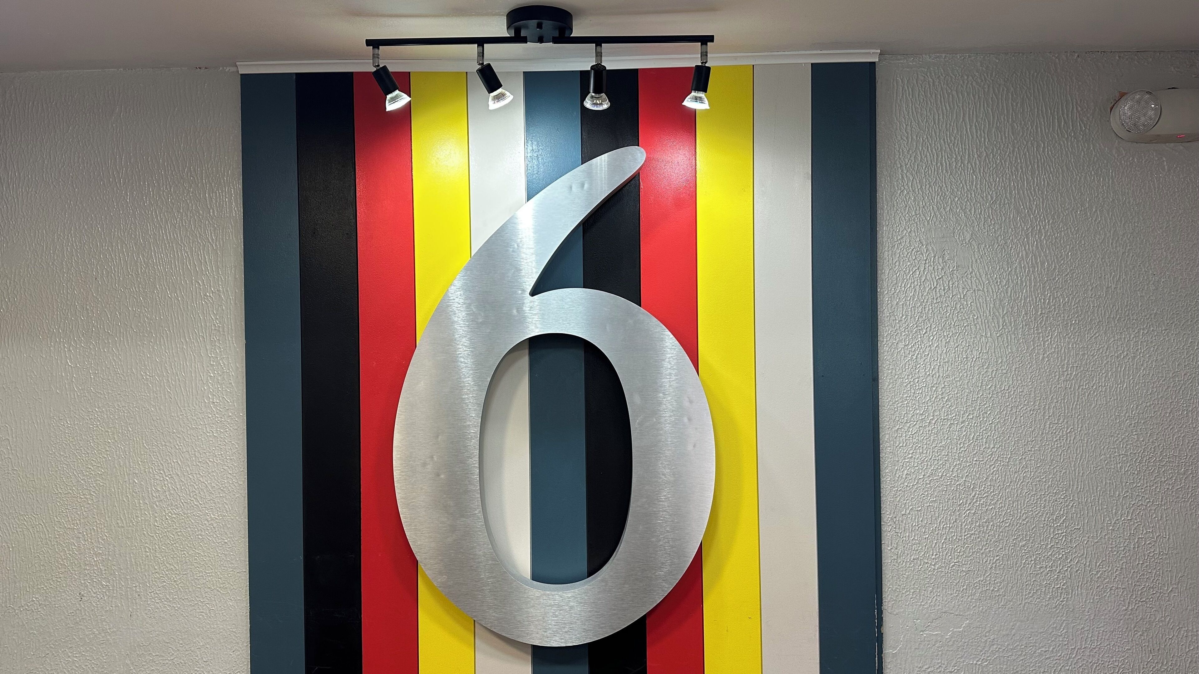 A large metallic number '6' mounted on a wall with vertical stripes in red, yellow, blue, and black.