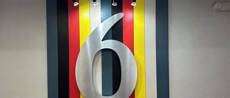 A large metallic number '6' mounted on a wall with vertical stripes in red, yellow, blue, and black.
