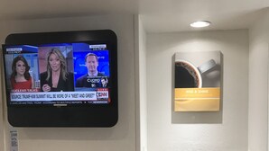 A television displaying a news program, a wall-mounted sign with a coffee cup, and a small seating area with cabinets. - Motel 6 Raleigh, NC - North (Raleigh)