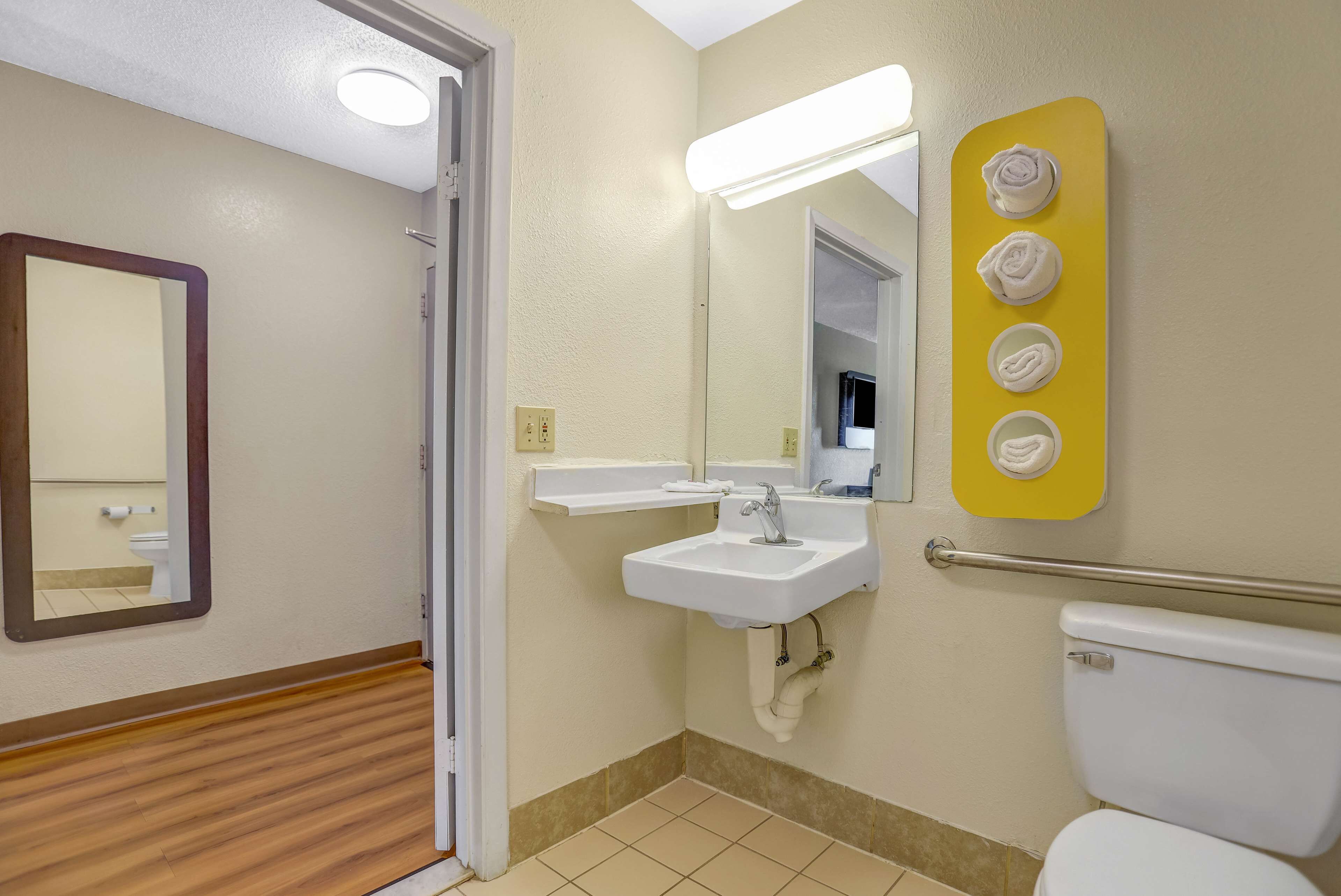 A bathroom with a wooden floor, a white sink, a mirror, a toilet, and a towel rack with four towels.