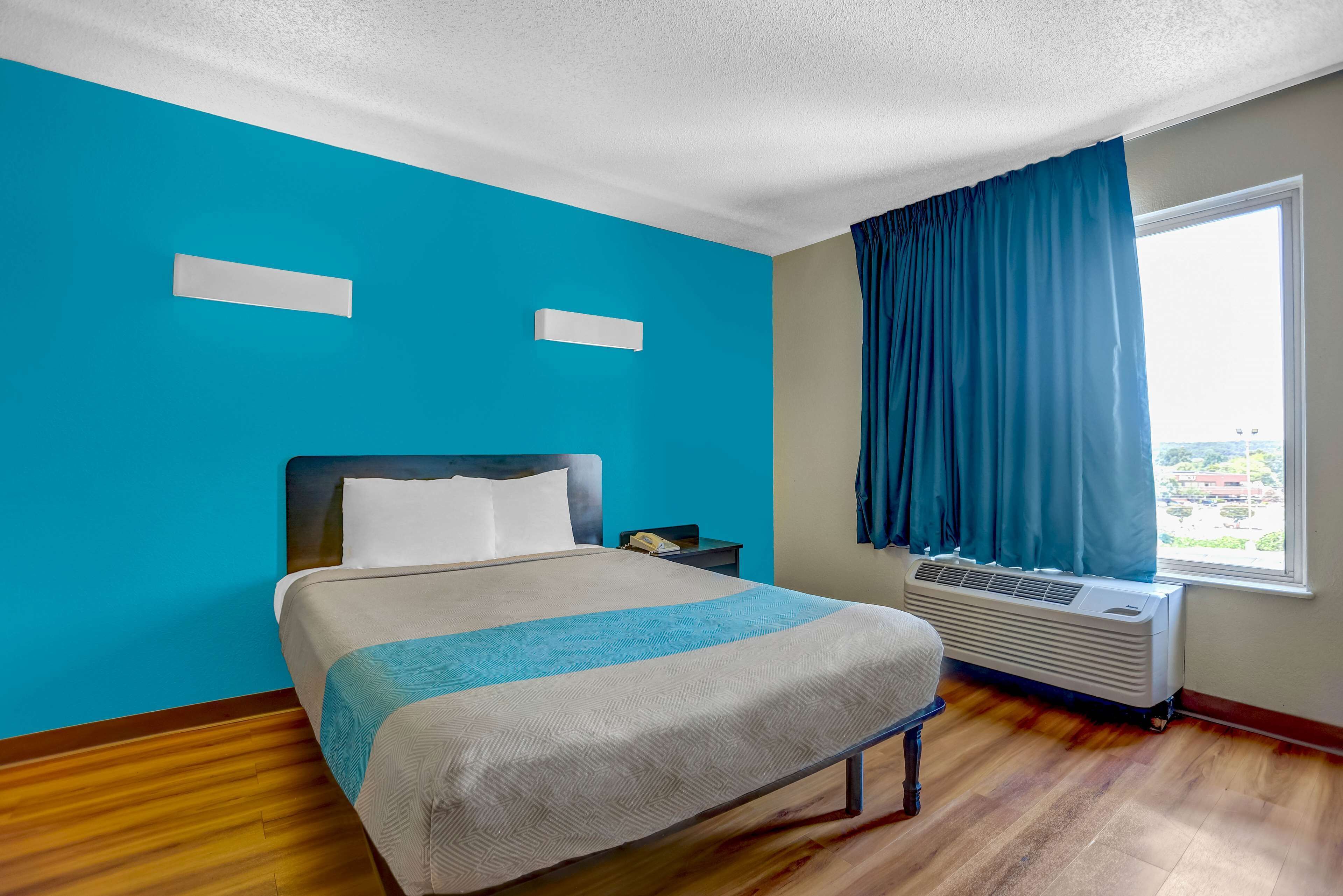 Una habitación de hotel con una cama, una mesita de noche, una ventana con cortinas y un equipo de aire acondicionado.