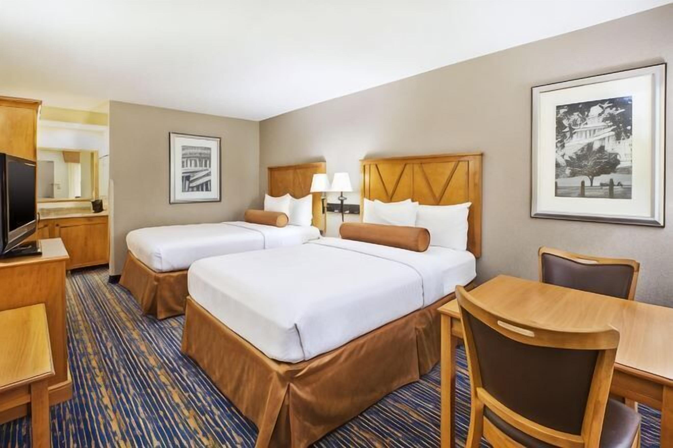 A hotel room with two beds, a desk, a chair, a TV, and framed pictures on the walls.