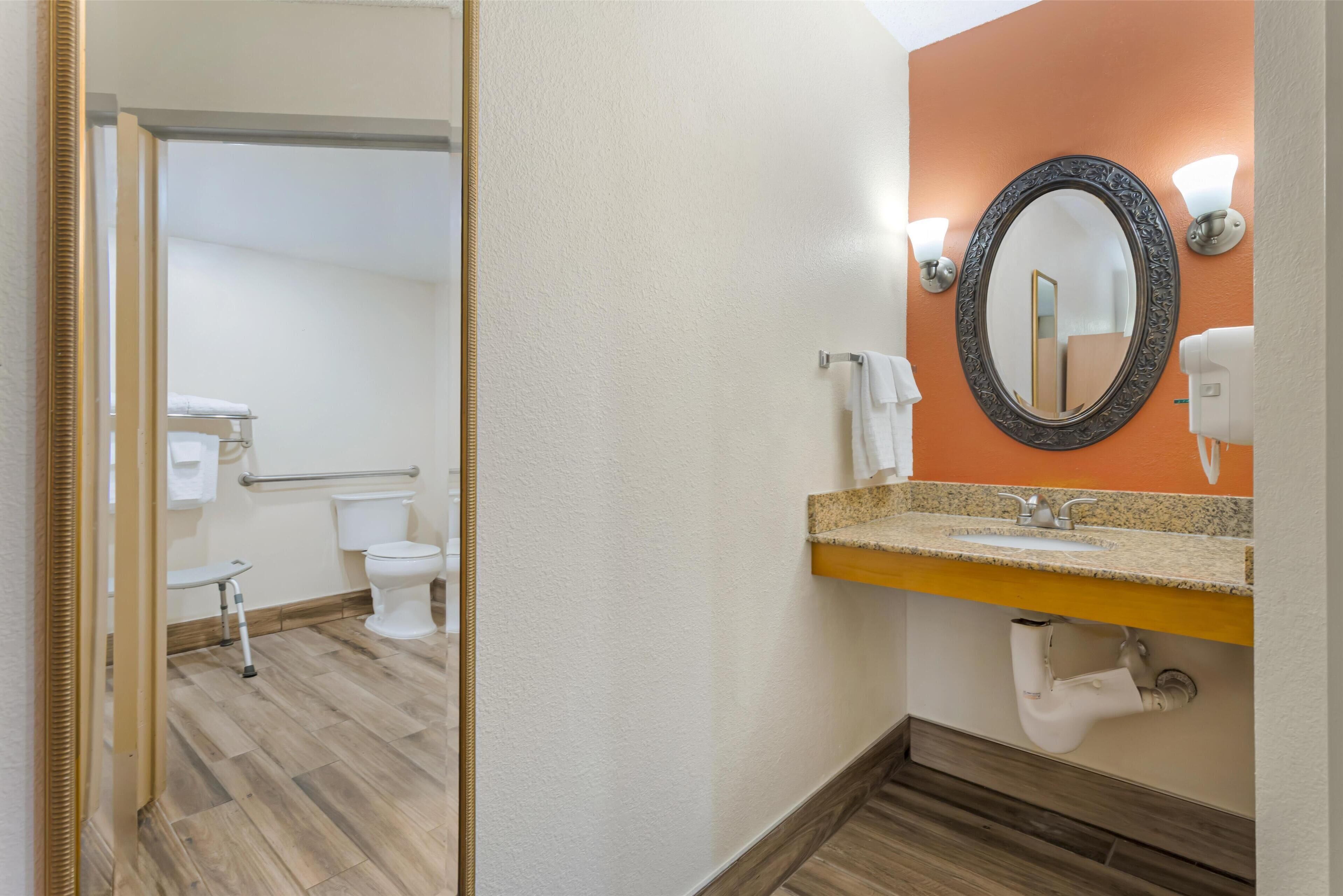 A bathroom with a wooden floor, a white toilet, a sink with a granite countertop, and a mirror with wall sconces.