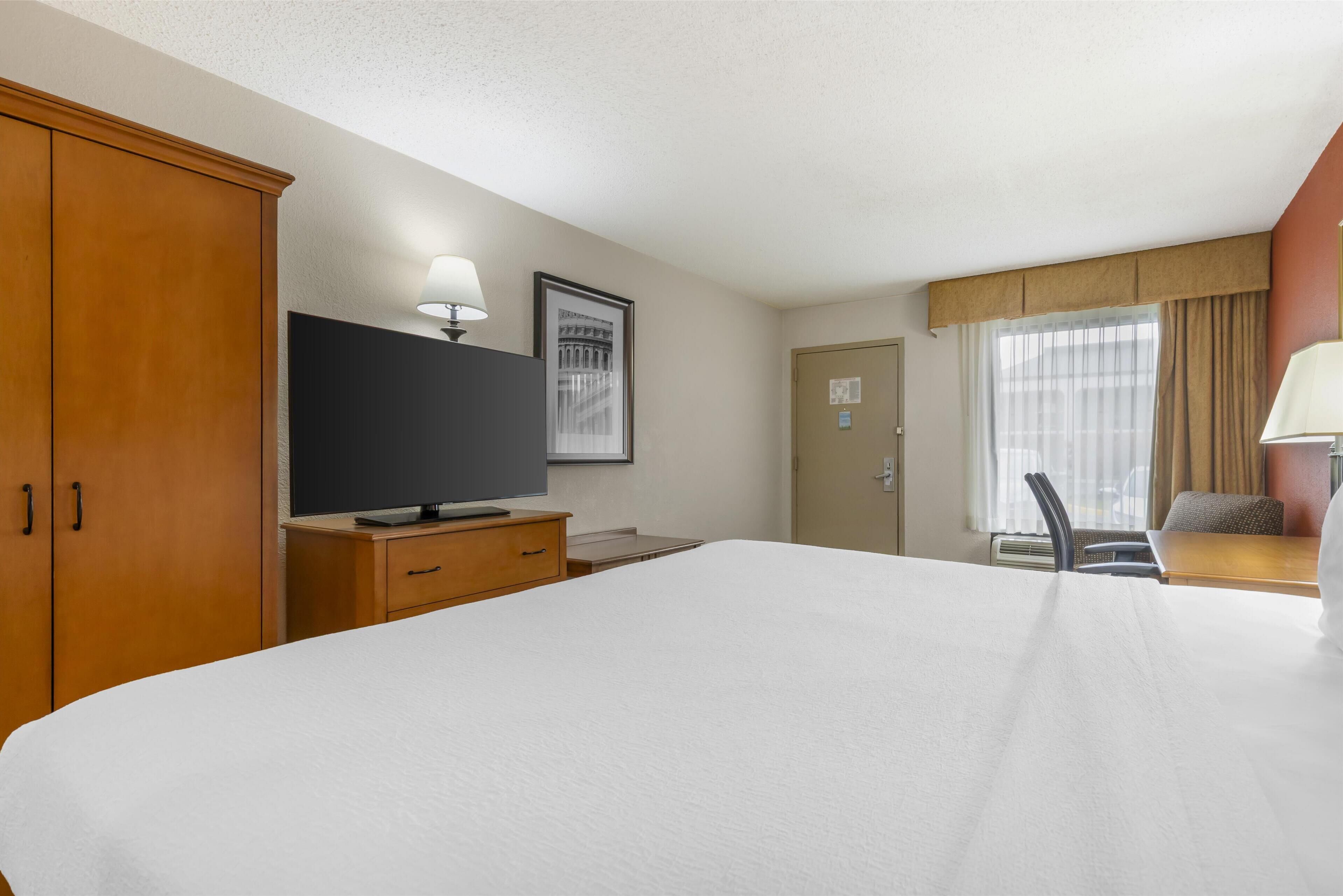 A hotel room with a large bed, a television, a desk with a lamp, and a window with curtains.
