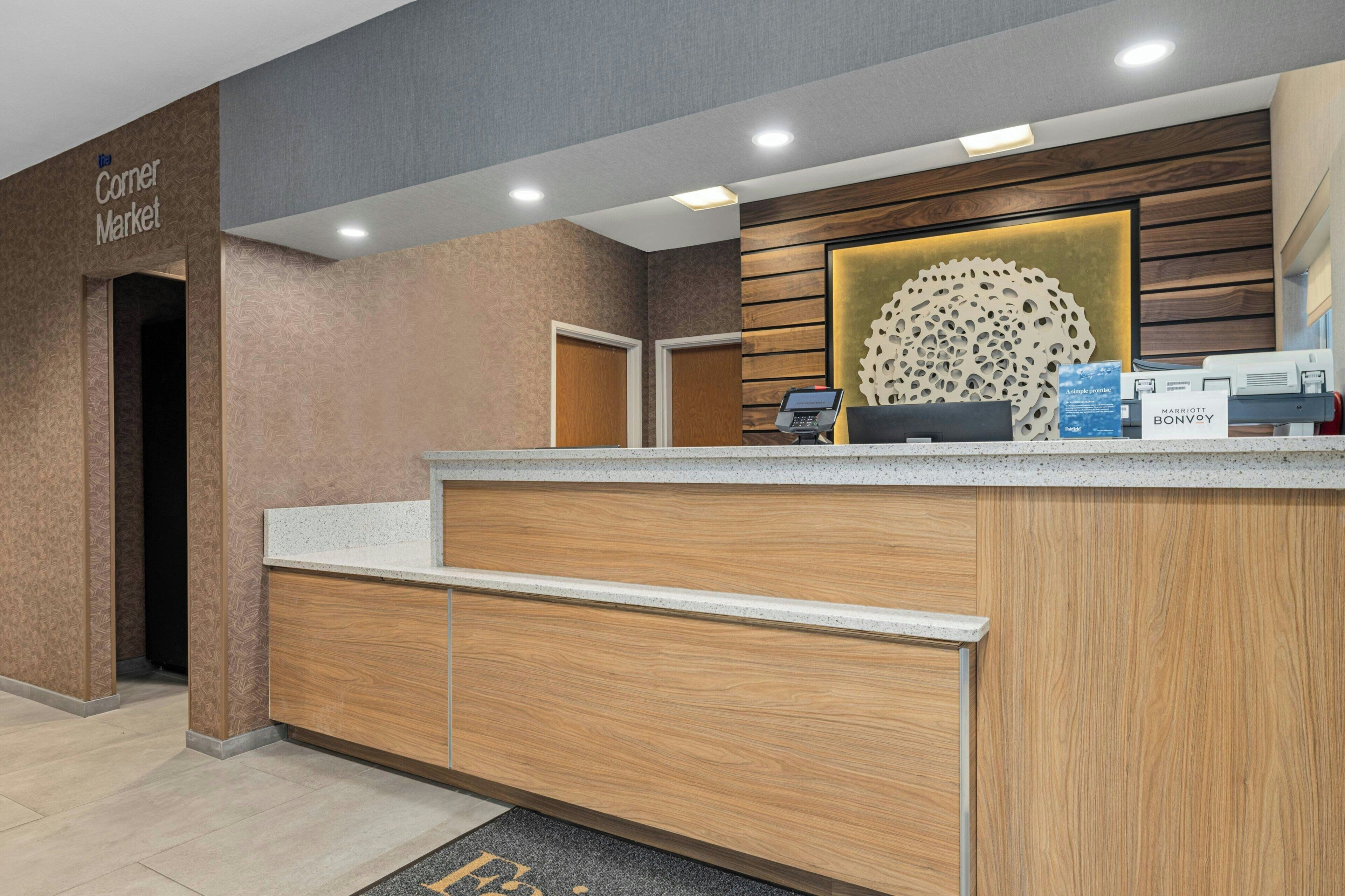 A hotel reception area with a wooden reception desk, a digital screen, and a sign for the Corner Market.