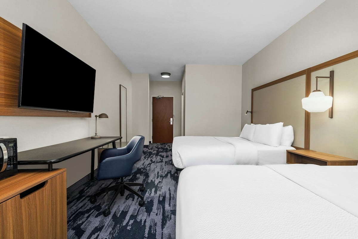 A hotel room with a flat-screen TV, a desk with a computer, a blue office chair, a bed, and a bedside table with a lamp.