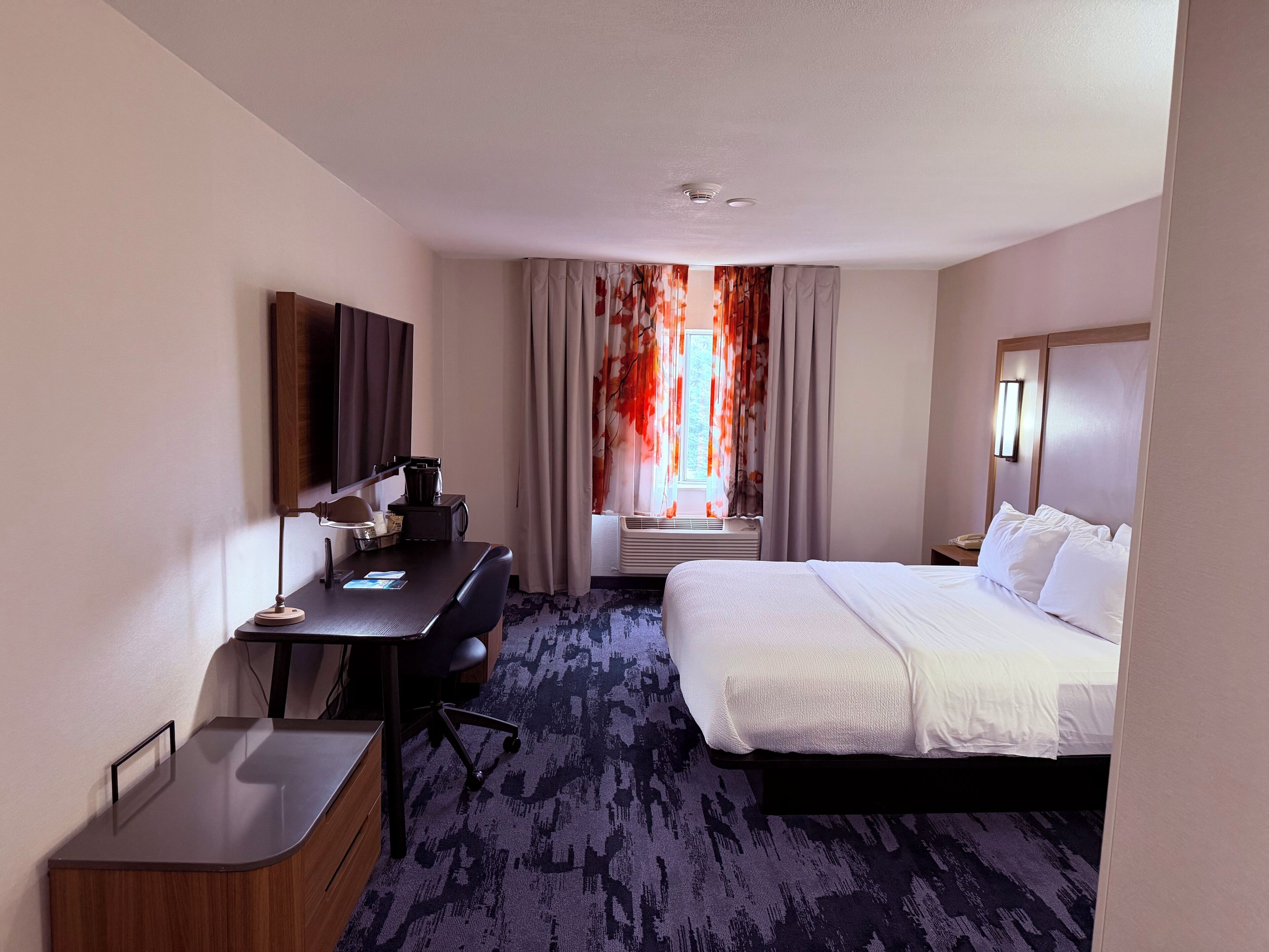 Quarto de hotel com cama, escrivaninha, cadeira, televisão e janela com cortinas.